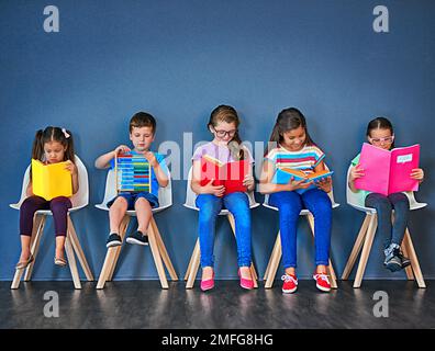 Restez curieux. Photo studio d'un groupe d'enfants assis sur des chaises et lisant des livres sur fond bleu. Banque D'Images