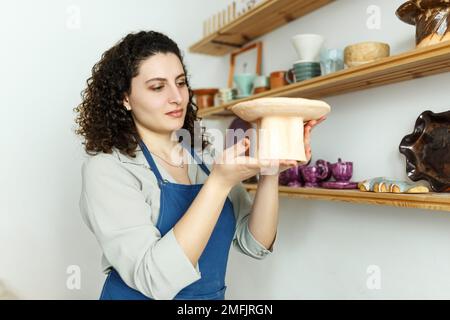 Jeune femme d'affaires dans son atelier est debout à côté de l'étagère avec des produits en céramique. Concept de démarrage de petites entreprises. Banque D'Images