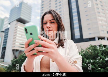 Gros plan portrait d'une fille de brune caucasienne regardant les nouvelles concentrées sur une application pour smartphone. Jeune femme caucasienne sérieuse utilisant un téléphone portable debout à l'extérieur. Photo de haute qualité Banque D'Images
