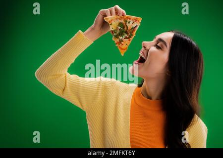 jeune femme en cavalier jaune tenant une délicieuse pizza près de la bouche ouverte isolée sur vert, image de stock Banque D'Images