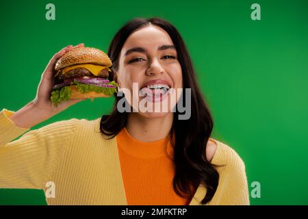 brunette femme tenant un hamburger savoureux et collant hors de la langue isolée sur vert, image de stock Banque D'Images