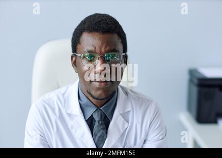 Portrait minimal de la vue avant d'un jeune médecin noir regardant l'appareil photo tout en étant assis au bureau Banque D'Images