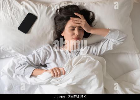 Une jeune femme européenne triste se réveille, se presse de la main à la tête, souffre de maux de tête, a de mauvais rêves Banque D'Images