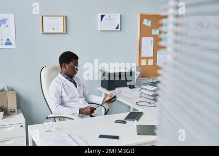 Portrait grand angle d'un jeune médecin noir qui se détend dans une chaise au bureau et lit des documents Banque D'Images