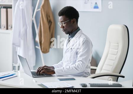 Portrait avec vue latérale minimale d'un jeune médecin noir utilisant un ordinateur portable au bureau et en tapant pendant son travail en clinique Banque D'Images