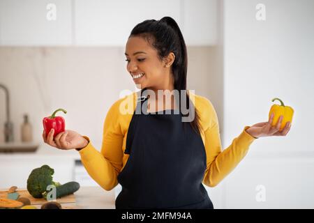 Joyeuse femme afro-américaine millénaire en tablier choisit des poivrons dans les mains à la table avec des légumes Banque D'Images