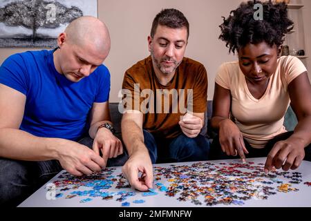 Des amis jouant des puzzles à la maison, sur une table blanche en bois. Rassembler les choses et résoudre les problèmes. Diversité et plaisir dans une amitié. Banque D'Images