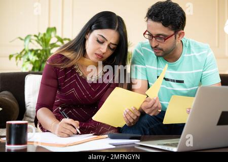 Un jeune couple indien sérieux calculant des dépenses ou des factures mensuelles devant un ordinateur portable tout en étant assis à la maison - concept d'investissement, budget et Banque D'Images