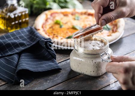 Les mains du cuisinier ouvrent un pot de levain de seigle, en arrière-plan une pizza faite de levain. Banque D'Images
