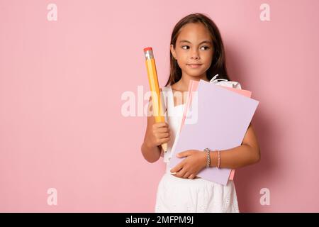Souriant actif excellent élève écofille tenant des livres et un gros crayon allant à l'école isolée dans fond rose Banque D'Images