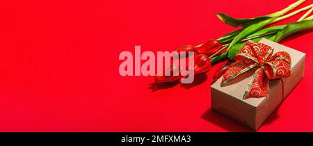Concept cadeau de la Saint-Valentin. Fond rouge romantique avec bouquet de tulipes lumineux et boîte surprise enveloppée. Lumière dure, ombre foncée, style minimaliste, Banque D'Images