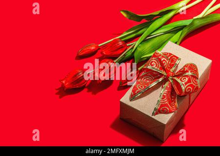 Concept cadeau de la Saint-Valentin. Fond rouge romantique avec bouquet de tulipes lumineux et boîte surprise enveloppée. Lumière dure, ombre foncée, style minimaliste, Banque D'Images