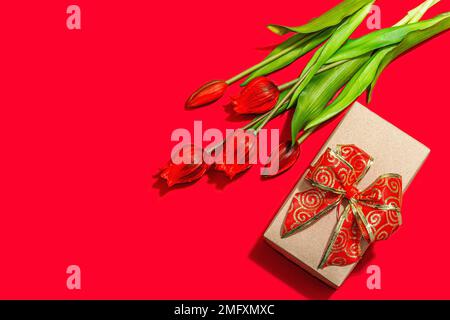 Concept cadeau de la Saint-Valentin. Fond rouge romantique avec bouquet de tulipes lumineux et boîte surprise enveloppée. Lumière dure, ombre foncée, style minimaliste, Banque D'Images