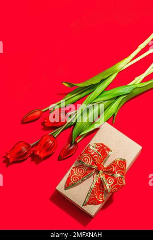 Concept cadeau de la Saint-Valentin. Fond rouge romantique avec bouquet de tulipes lumineux et boîte surprise enveloppée. Lumière dure, ombre foncée, style minimaliste, Banque D'Images