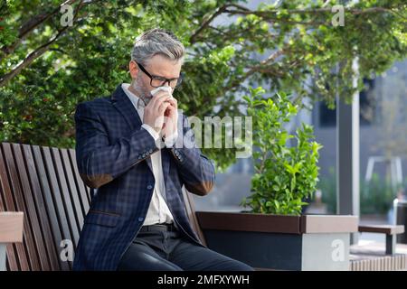 Homme d'affaires mature travaillant à l'extérieur avec un ordinateur portable, homme âgé éternuant des allergies, assis sur le banc du parc par beau soleil, patron en costume d'affaires. Banque D'Images