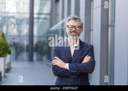 Portrait d'un homme d'affaires mûr aux cheveux gris, d'un patron senior souriant et regardant un appareil photo aux bras croisés, d'un homme aux cheveux gris à l'extérieur du bâtiment de bureau, d'un investisseur et d'un propriétaire prospères en costume d'affaires. Banque D'Images