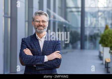 Portrait d'un homme d'affaires mûr aux cheveux gris, d'un patron senior souriant et regardant un appareil photo aux bras croisés, d'un homme aux cheveux gris à l'extérieur du bâtiment de bureau, d'un investisseur et d'un propriétaire prospères en costume d'affaires. Banque D'Images