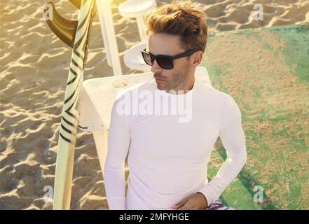 Un jeune homme souriant est assis sur une plage à côté de sa planche de surf, portant des lunettes de soleil sombres et une chemise blanche vierge à manches longues en lycra. Maquette verticale- Banque D'Images