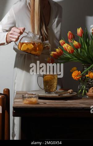 Femme versant le thé de la théière dans la théière. Thé du matin dans la cuisine sur une table décorée de fleurs de tulipe. Banque D'Images