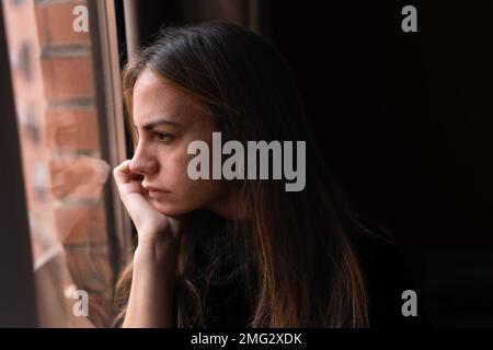 Vue latérale d'une jeune femme attentionnés avec de longs cheveux foncés penchés au menton sur la main et donnant sur la fenêtre à la maison Banque D'Images