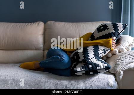 Corps complet de femme mélancolique non reconnaissable dans des vêtements décontractés couvrant le visage avec un oreiller tout en étant allongé sur un canapé confortable dans l'appartement Banque D'Images