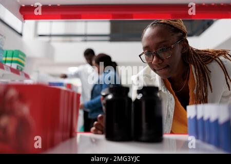 Femme pharmacien mettant des flacons de pilules sur la tablette de pharmacie, gros plan sélectif foyer. Vendeur afro-américain prenant des paquets de comprimés, service pharmaceutique, vente de médicaments Banque D'Images