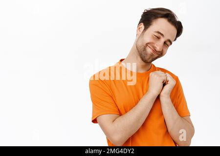 Un adorable homme souriant tient les mains sur le cœur et se réjouit, se tient dans un t-shirt orange Banque D'Images