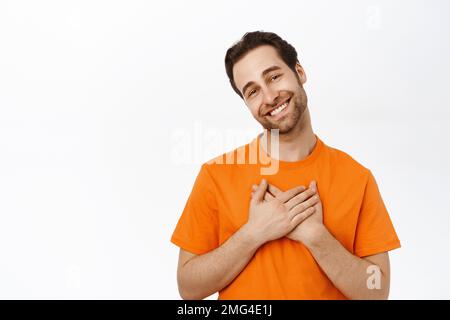 Un adorable homme souriant tient les mains sur le cœur et se réjouit, se tient dans un t-shirt orange Banque D'Images