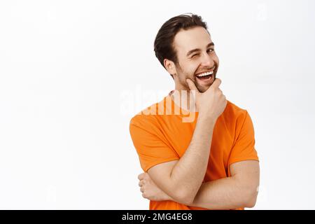 Image d'un charmant homme de brunette avec barbe, se tordre et sourire à l'appareil photo avec visage heureux, debout sur fond blanc Banque D'Images