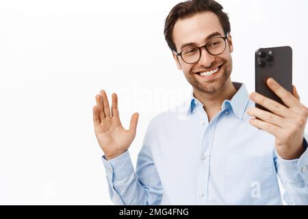 Beau homme d'affaires qui fait la démonstration de smth sur appel vidéo. Un employé de bureau qui parle sur un téléphone portable, agite la main, dit bonjour, fond blanc Banque D'Images