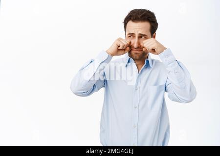 Homme adulte pleurant et essuyant ses larmes, ayant l'air triste ou offensé, en boussant, debout sur fond blanc Banque D'Images