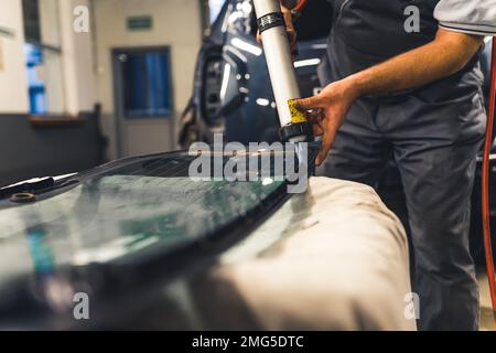 Homme méconnaissable appliquant de l'adhésif industriel sur la vitre avant de l'installer dans la voiture. Travaux de garage. Se concentrer sur l'arrière-plan. Prise de vue horizontale en intérieur. Photo de haute qualité Banque D'Images