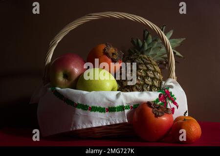 La vie d'un panier en osier avec des fruits assortis avec un ananas, des persimmons, des mandarines et des pommes sur un fond sombre et un tapis rouge Banque D'Images