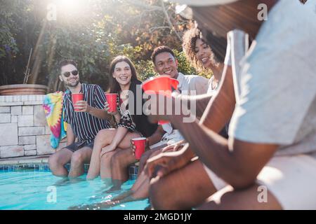 Vacances, boissons et amis parlant dans une piscine lors d'une fête d'été, d'une fête ou d'un événement à la maison. La diversité, heureux et les gens parlent, avoir plaisir et Banque D'Images