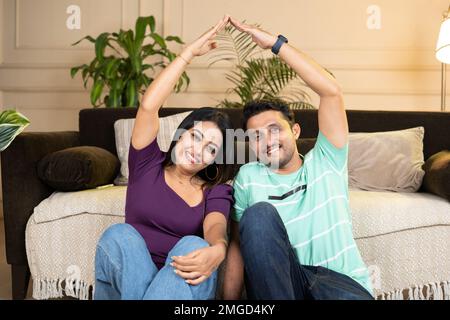 un couple souriant se montre heureux en faisant un geste sur le toit de la maison en joignant les mains tout en regardant l'appareil photo là-bas nouvelle maison ou appartement - concept de nouvelle maison Banque D'Images