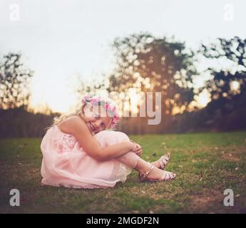 Je ne veux pas que ce jour finisse. Une petite fille heureuse assise sur le sol et regardant la caméra à l'extérieur dans la nature. Banque D'Images