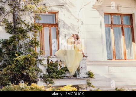 une belle femme souriante et gentille dans une magnifique robe jaune se dresse sur le balcon d'une ancienne maison d'époque Banque D'Images