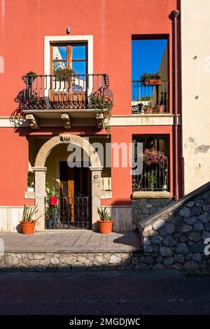 Façade d'une maison rouge avec porte, balcon, trois fenêtres et fleurs en pots dans l'une des ruelles de la ville de Milazzo. Banque D'Images