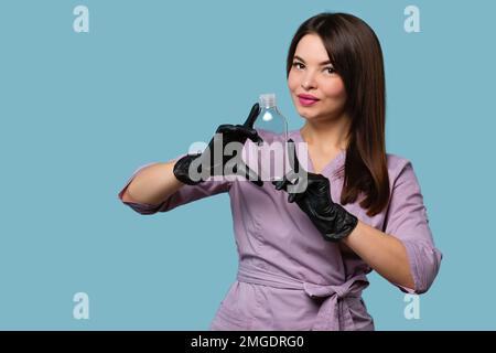 Une esthéticienne peut contenir une bouteille en verre transparent vide. Femme esthéticienne en uniforme rose et gants noirs sur fond de studio bleu clair Banque D'Images