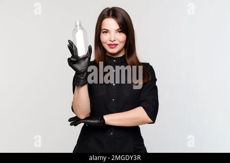 Une esthéticienne peut contenir une bouteille en verre transparent vide. Femme esthéticienne en uniforme noir et gants sur fond blanc de studio Banque D'Images