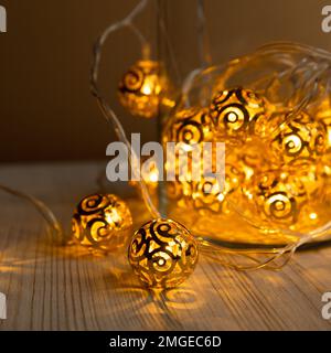 La guirlande du nouvel an. Boules métalliques dorées dans un pot en verre sur fond de table en bois clair. Guirlande lumineuse de Noël jaune or avec ampoules LED. Vue avant. Banque D'Images