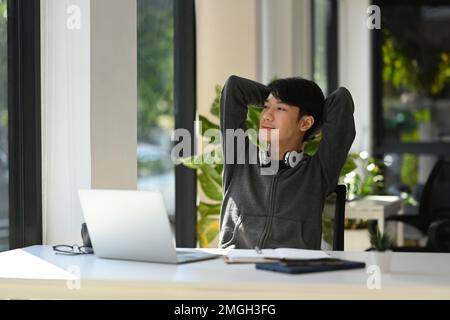 Jeune homme asiatique détendu travailleur assis dans une chaise de bureau avec les mains clastées derrière la tête et regardant par la fenêtre Banque D'Images