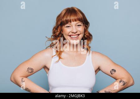 Portrait d'une jeune femme heureuse avec des tatouages de bras debout dans un studio. Jeune femme gaie avec des cheveux de gingembre debout contre un fond bleu dans un W Banque D'Images