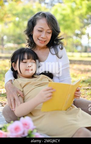 Petite-fille asiatique mignonne à poser sur sa grand-mère, à lire un livre ou à écouter une histoire assez racontée par sa grand-mère tout en pique-niquant dans le parc Banque D'Images