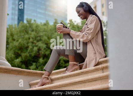 Casque audio, téléphone et femme noire avec café tout en étant assis sur les marches de la ville. Technologie, pause thé et une femme heureuse avec de la caféine et Banque D'Images