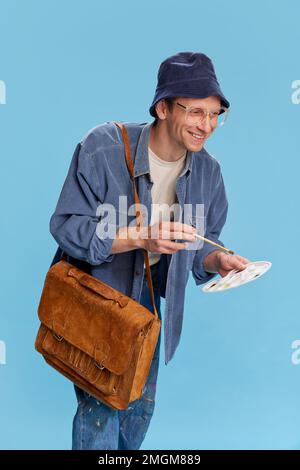 Choisir une couleur pour une photo future. Portrait d'un jeune homme en jeans, peintre, artiste posant sur fond bleu studio. Concept des émotions Banque D'Images