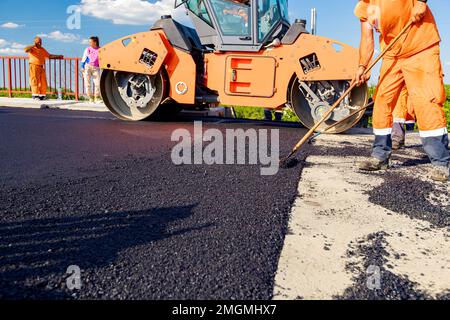 Les travailleurs utilisent des râteaux pour niveler, mettre en place une couche de tarmac frais à de bonnes mesures, le rouleau vapeur est flatter de l'asphalte frais . Banque D'Images