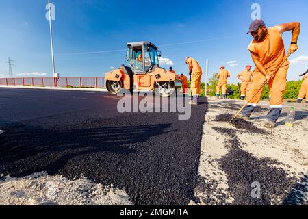 Zrenjanin, Voïvodine, Serbie - 8 juin 2021: Les travailleurs utilisent des râteaux pour niveler, mettre en place une couche de tarmac frais à de bonnes mesures, le rouleau vapeur est en train de flatter Banque D'Images