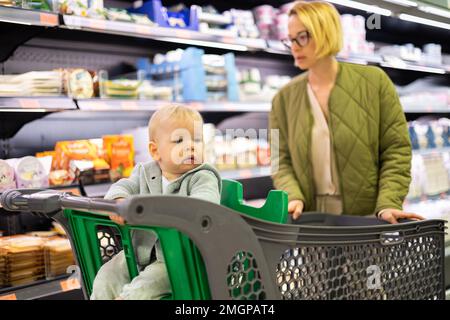 Casualy mère habillée choisissant des produits en conserve dans le rayon de supermarché épicerie avec son bébé garçon enfant dans poussette Banque D'Images