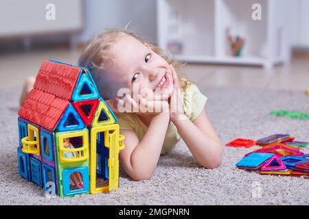 Jolie fille curly à côté d'une maison faite d'un constructeur magnétique à la maison sur le tapis dans la chambre des enfants. Banque D'Images